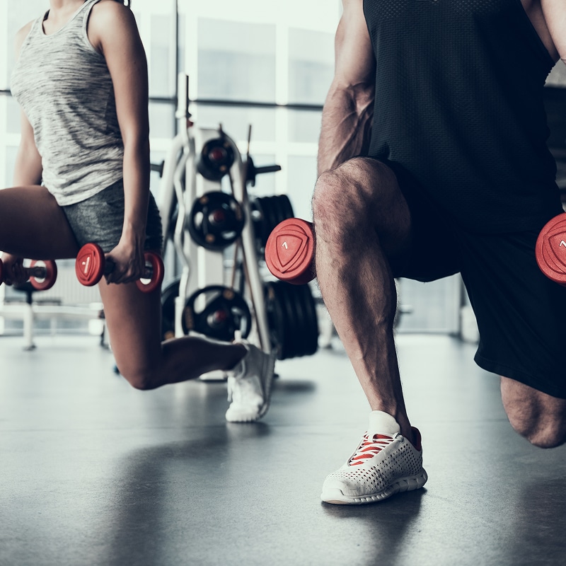 Couple musclé faisant des fentes avec haltères rouges (poids 1) dans une salle de sport moderne, concentrés sur l'entraînement des jambes.