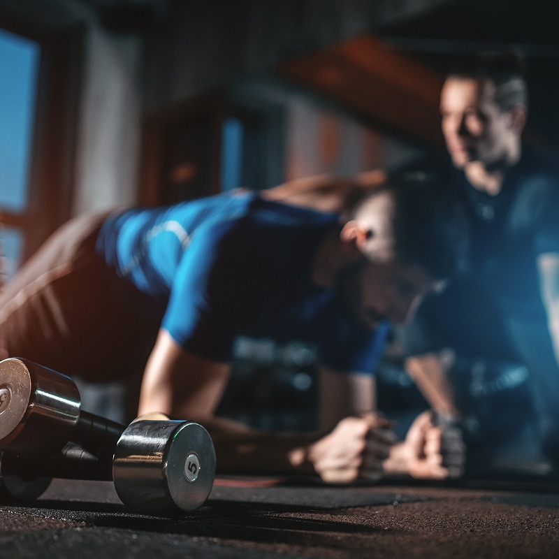 Homme en T-shirt bleu effectuant une planche sur un tapis avec des haltères de 5 kg au premier plan, coach observant.