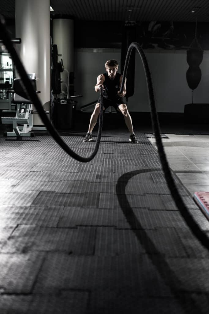 Homme musclé en tenue de sport noire utilisant des cordes de combat (battle ropes) dans une salle de sport sombre et texturée.