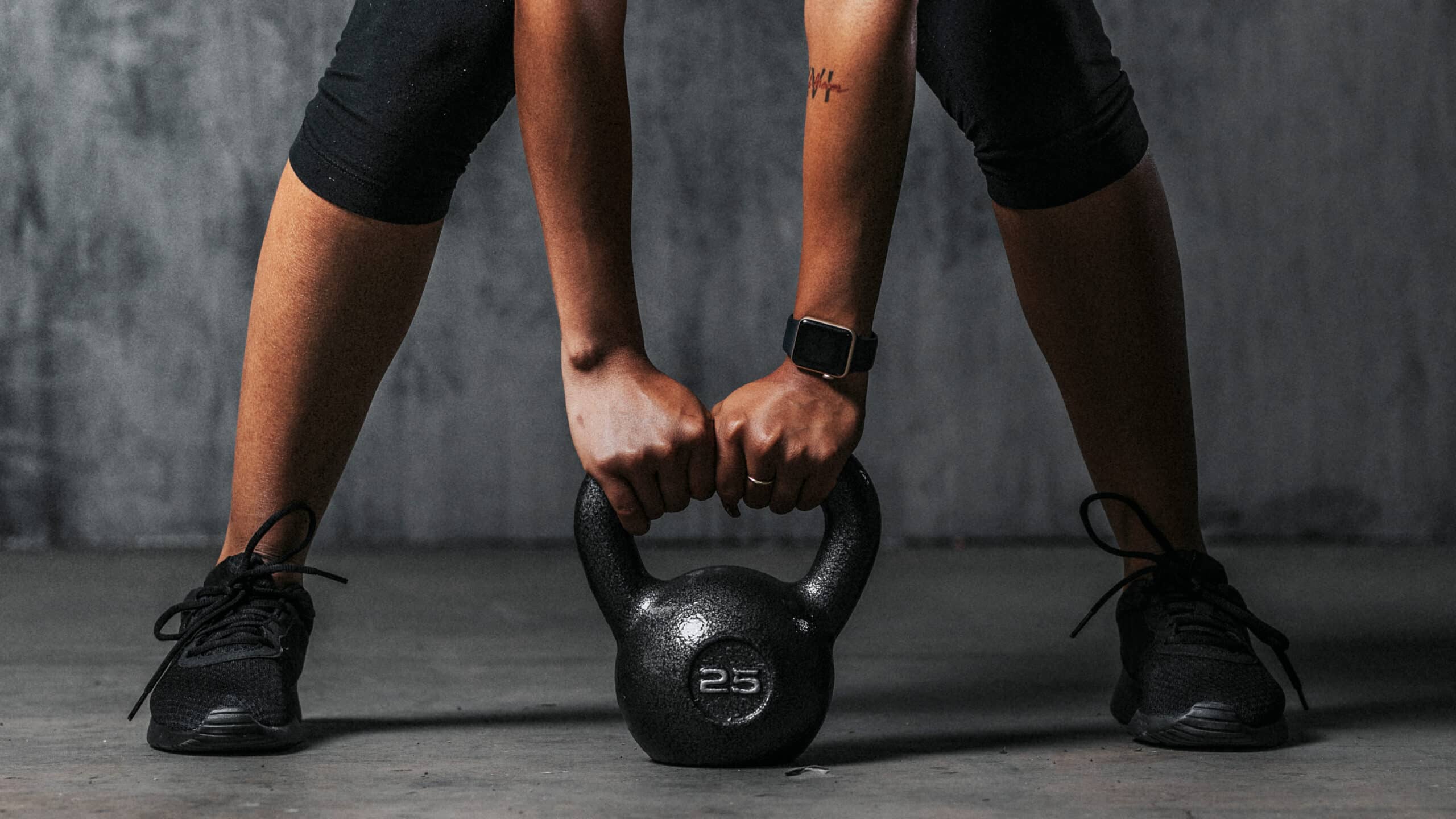 Entraînement avec kettlebell de 25, personne avec montre connectée, jambes écartées, tenant l'équipement sur sol gris.