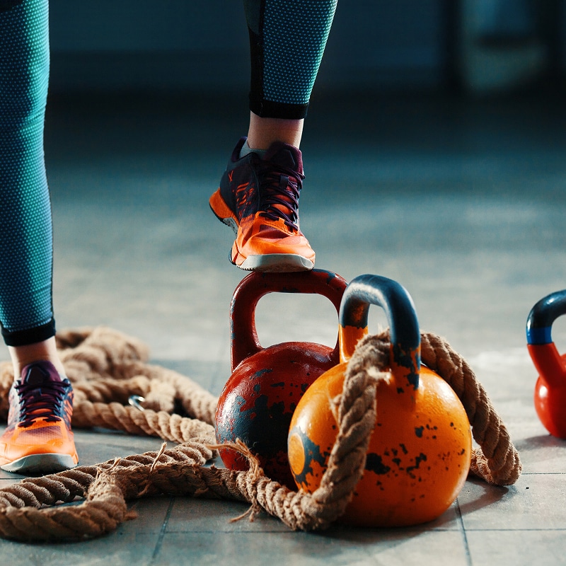 Gros plan sur des jambes vêtues de leggings bleus et de baskets orange, un pied posé sur une kettlebell rouge lors d'un entraînement de fitness avec des cordes.