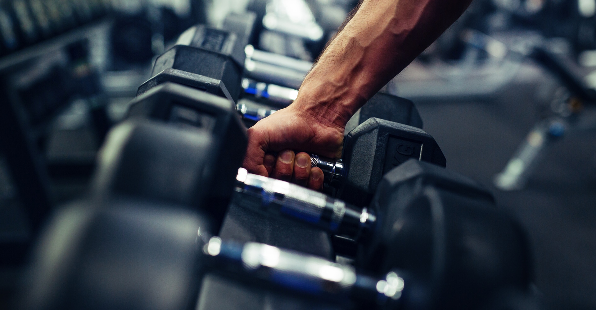 Gros plan sur une main musclée saisissant fermement l'haltère chromé d'un rack dans une salle de sport sombre et faiblement éclairée.