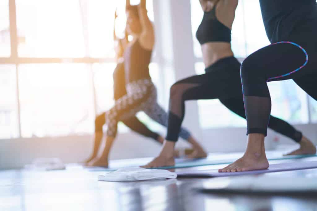 Femmes pratiquant le yoga en posture du guerrier (Virabhadrasana) dans une salle lumineuse, plan centré sur les jambes et les pieds.
