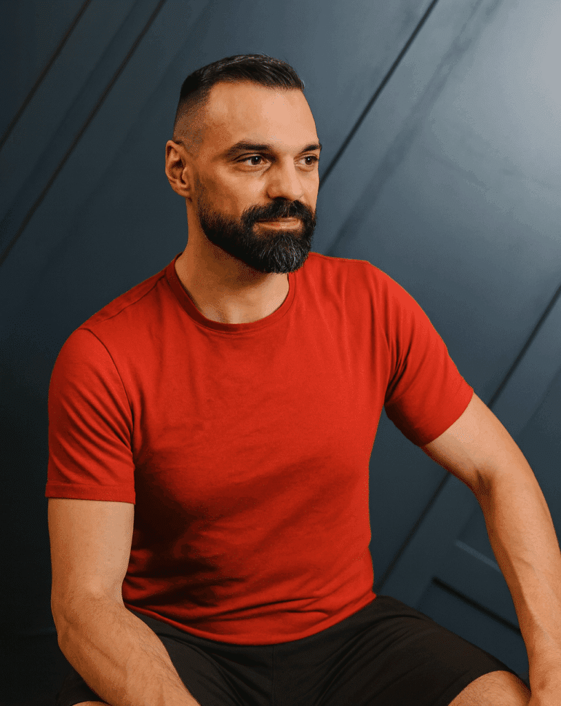Homme barbu au look soigné, vêtu d'un t-shirt rouge vif et d'un short noir, posant sur un fond bleu foncé texturé, regardant au loin avec sérieux.