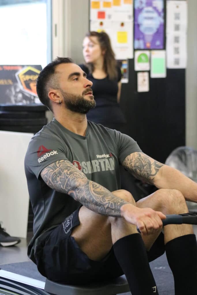 Homme barbu aux bras tatoués s'entraînant sur un rameur Reebok Spartan, concentré, une femme est floue en arrière-plan.