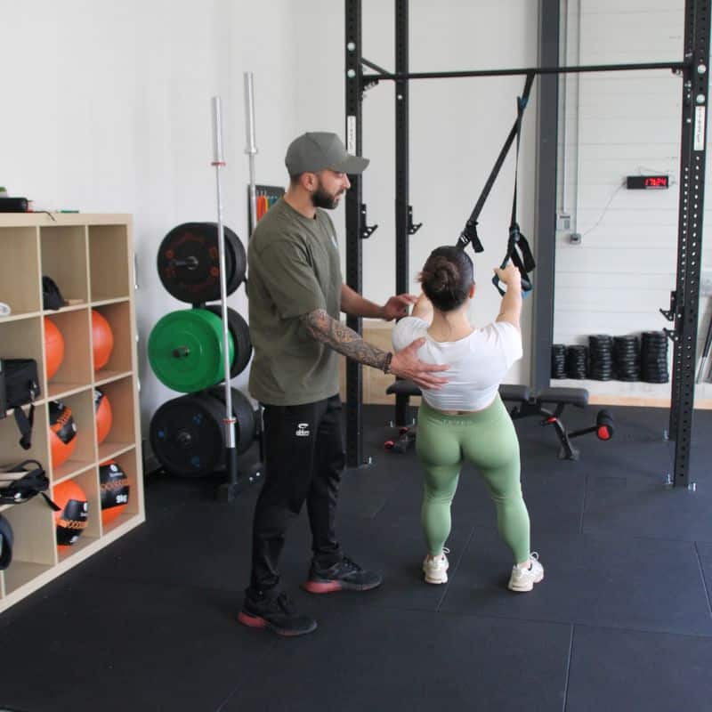 Coach Sportif : Séance Personnalisée de Musculation Coach sportif guidant une femme en plein exercice de suspension TRX dans une salle de musculation équipée.