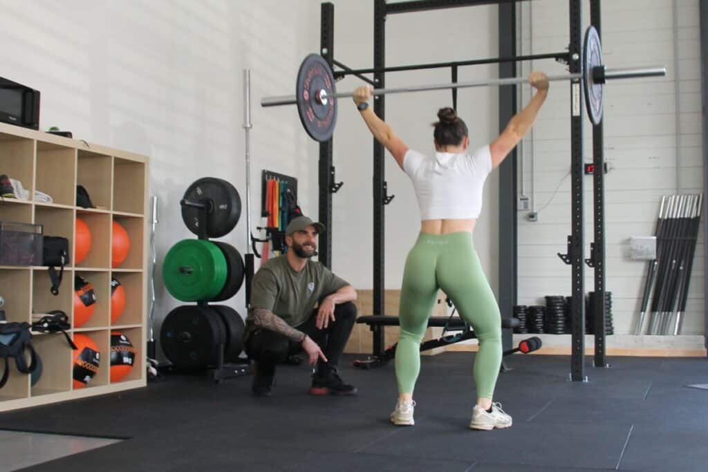 Haltérophilie et musculation : coaching personnalisé femme Femme soulevant une barre d'haltérophilie sous l'œil attentif d'un coach sportif dans une salle de musculation.