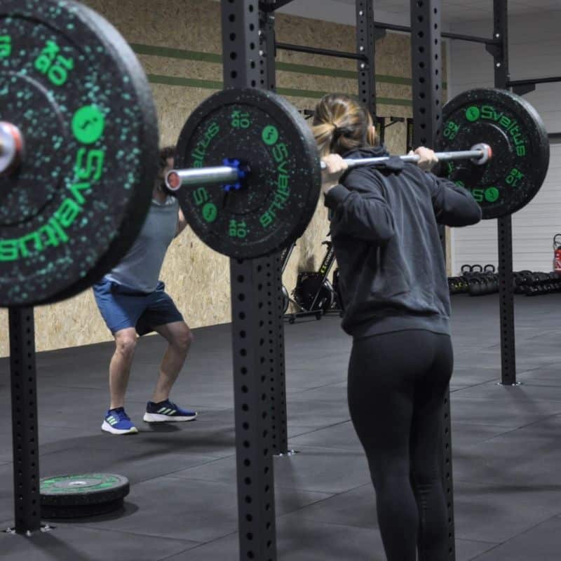 Entraînement Squat avec Barre et Poids Sveltus en Salle Femme effectuant un squat avec une barre Sveltus 10kg dans un rack de musculation moderne.